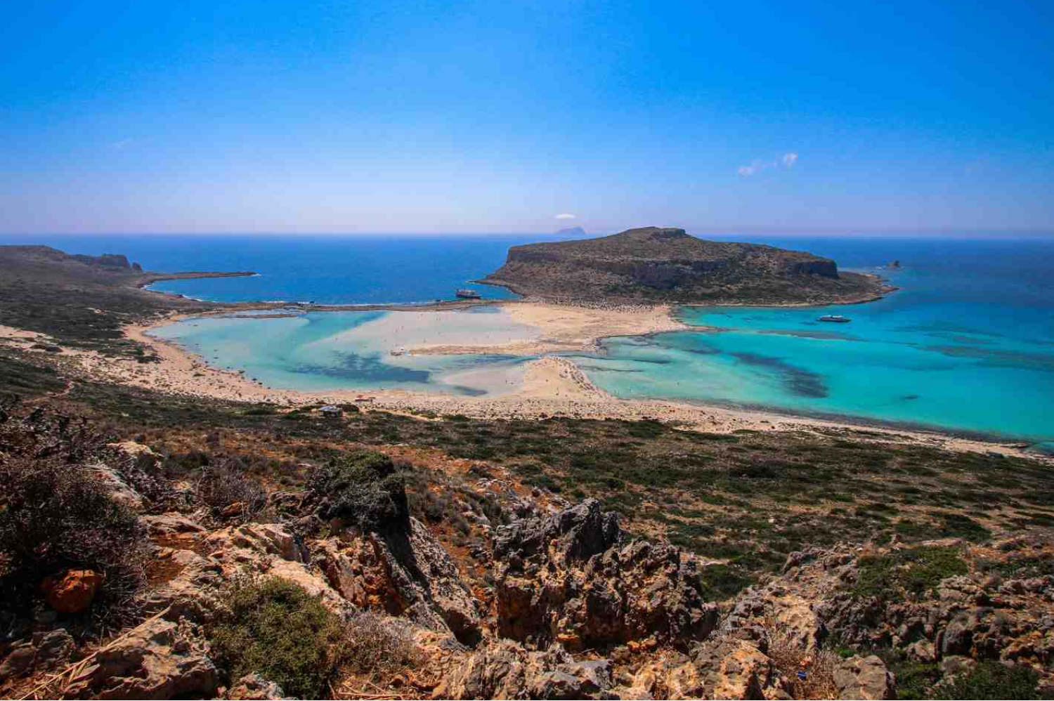 Vista panorâmica com mar azul, areia clara e montanhas ao fundo.
