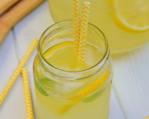 Limonada servida em pote de vidro com rodelas de limão, manjericão e canudos amarelos.